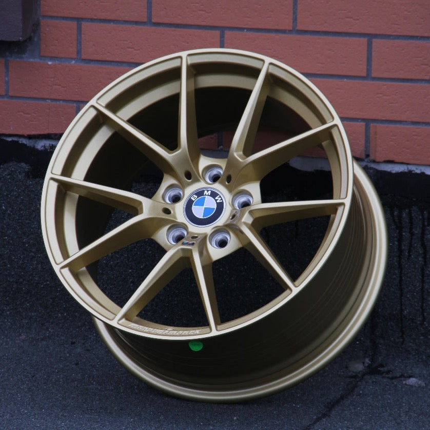 A gold BMW alloy wheel rests against a brick wall on a rough black surface. Center logo reads "BMW." "Competition" and a small green sticker are visible on the rim.