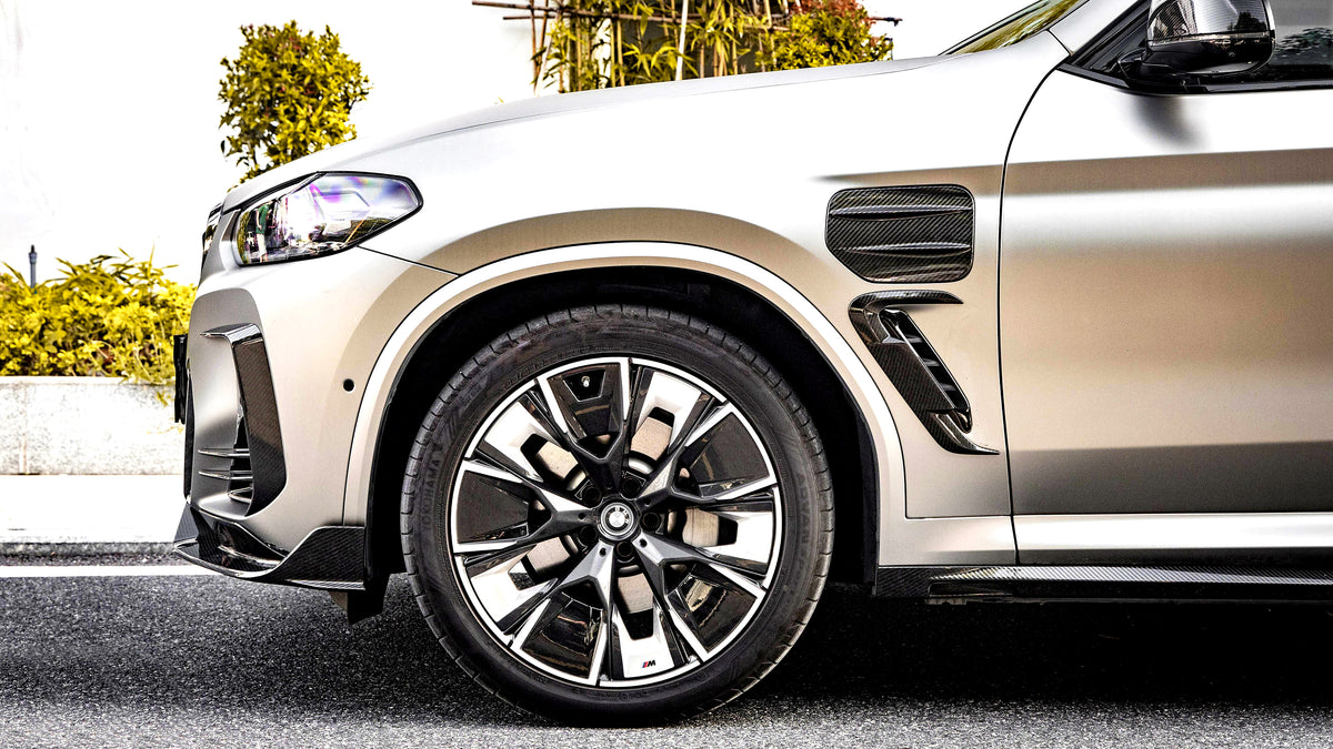 A silver car's front tire, featuring intricate alloy wheel design, is parked on a street. Greenery lines the background, providing a natural contrast to the vehicle.