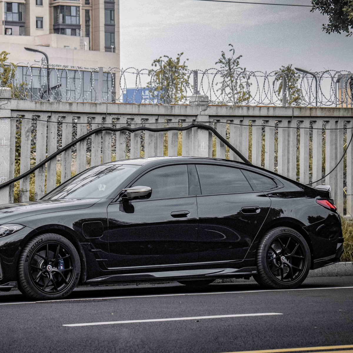 A black car is parked on a street with barbed wire atop a concrete fence. Apartment buildings and trees are visible in the background.