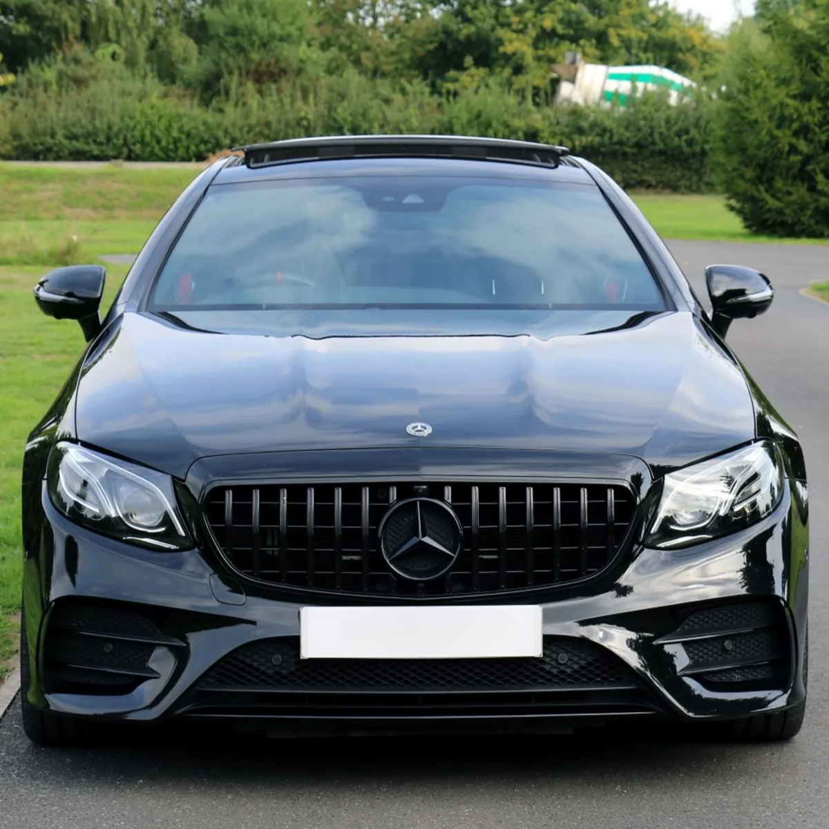 Car stationary on a road; glossy black body, wide grille with a logo, surrounded by lush green trees and grass. License plate is blank.