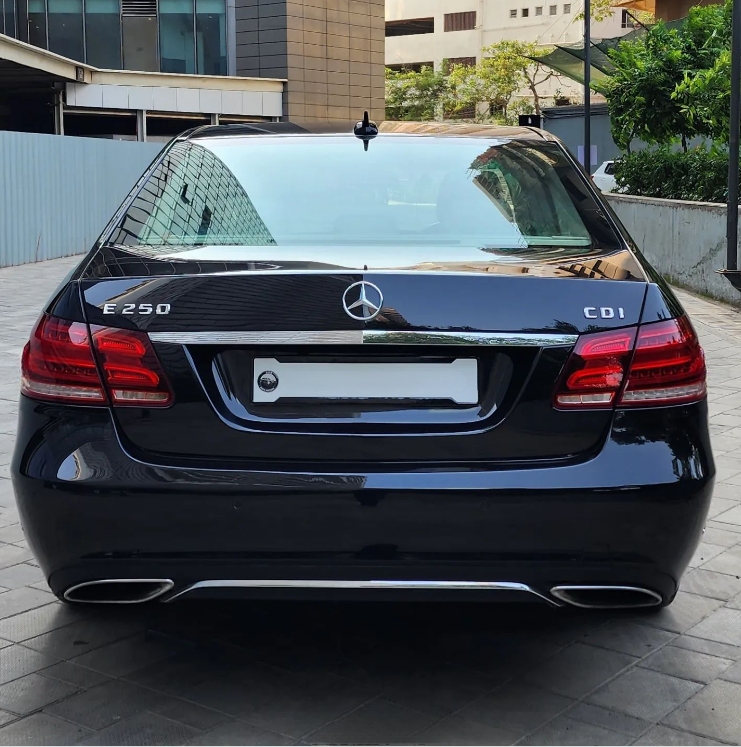 A black Mercedes-Benz E250 CDI is parked on a paved surface between modern buildings, with "E250" and "CDI" marked on the rear, featuring tinted windows and visible tail lights.