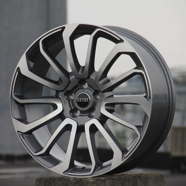 A modern alloy wheel stands upright, displaying intricate split-spoke design. Center hub bearing the text "RANGE ROVER." Positioned in an outdoor, urban setting with a concrete base.