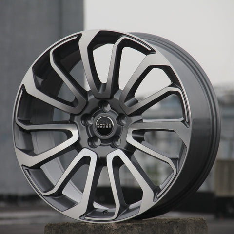 A modern alloy wheel stands upright, displaying intricate split-spoke design. Center hub bearing the text "RANGE ROVER." Positioned in an outdoor, urban setting with a concrete base.