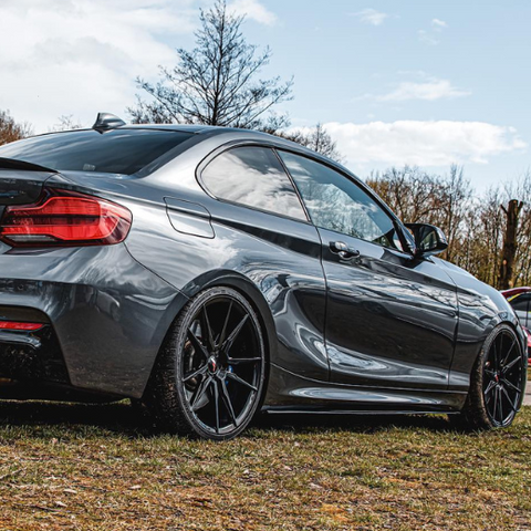 A sleek, metallic gray sports coupe stands parked on grassy terrain surrounded by trees under a partly cloudy sky. The car has dark alloy wheels and tinted windows.