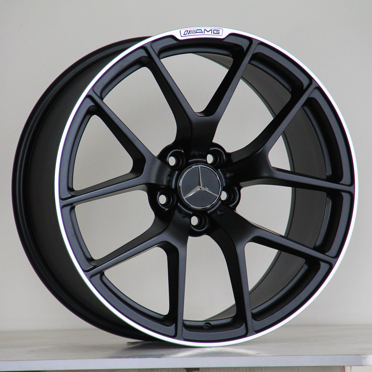 A black alloy wheel with a silver Mercedes logo, features the text "AMG" above the rim. It rests on a light surface against a plain background.