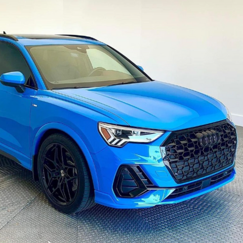 A bright blue SUV, featuring a prominent black grille and sleek headlights, is parked indoors on a checkered metal floor, surrounded by white walls.