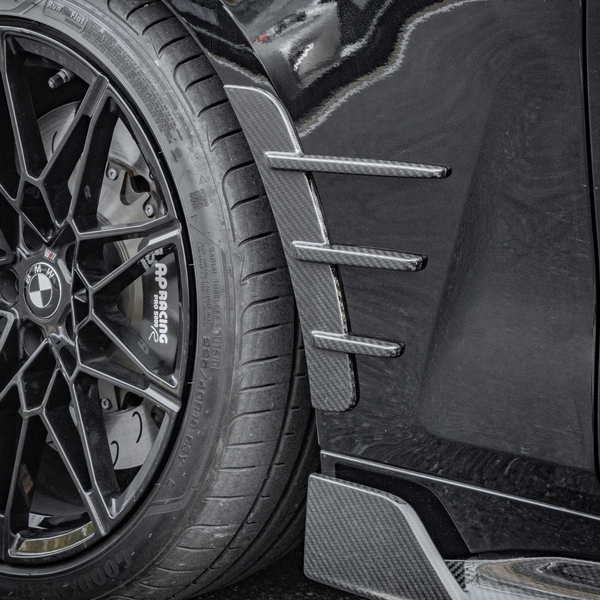A black BMW wheel with a multi-spoke design, featuring "AP Racing" brakes, is adjacent to carbon fiber detailing on a car body panel, enhancing aerodynamics on a smooth black surface.