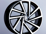 A polished, black-and-silver car wheel rim is displayed upright, featuring a VW logo at its center, against a softly lit, plain gray background.