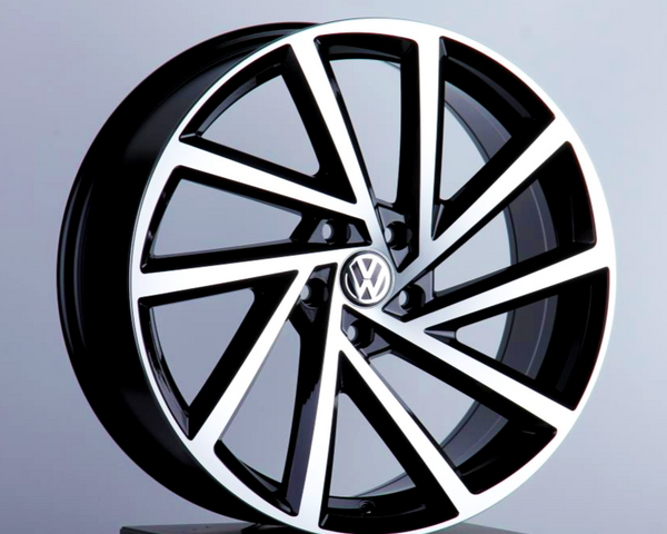 A polished, black-and-silver car wheel rim is displayed upright, featuring a VW logo at its center, against a softly lit, plain gray background.