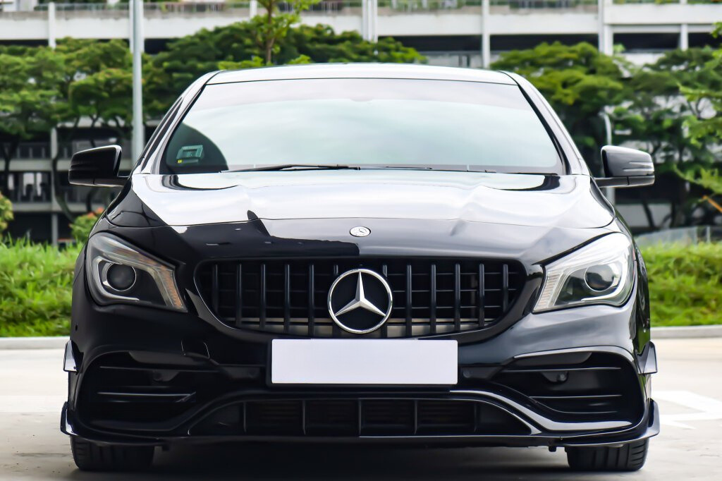A black Mercedes-Benz car is parked outdoors, facing forward. The background consists of greenery and a multi-story building.