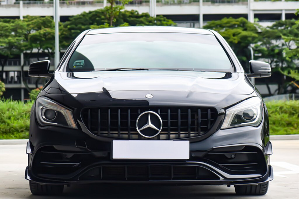 A black Mercedes-Benz car is parked outdoors, facing forward. The background consists of greenery and a multi-story building.