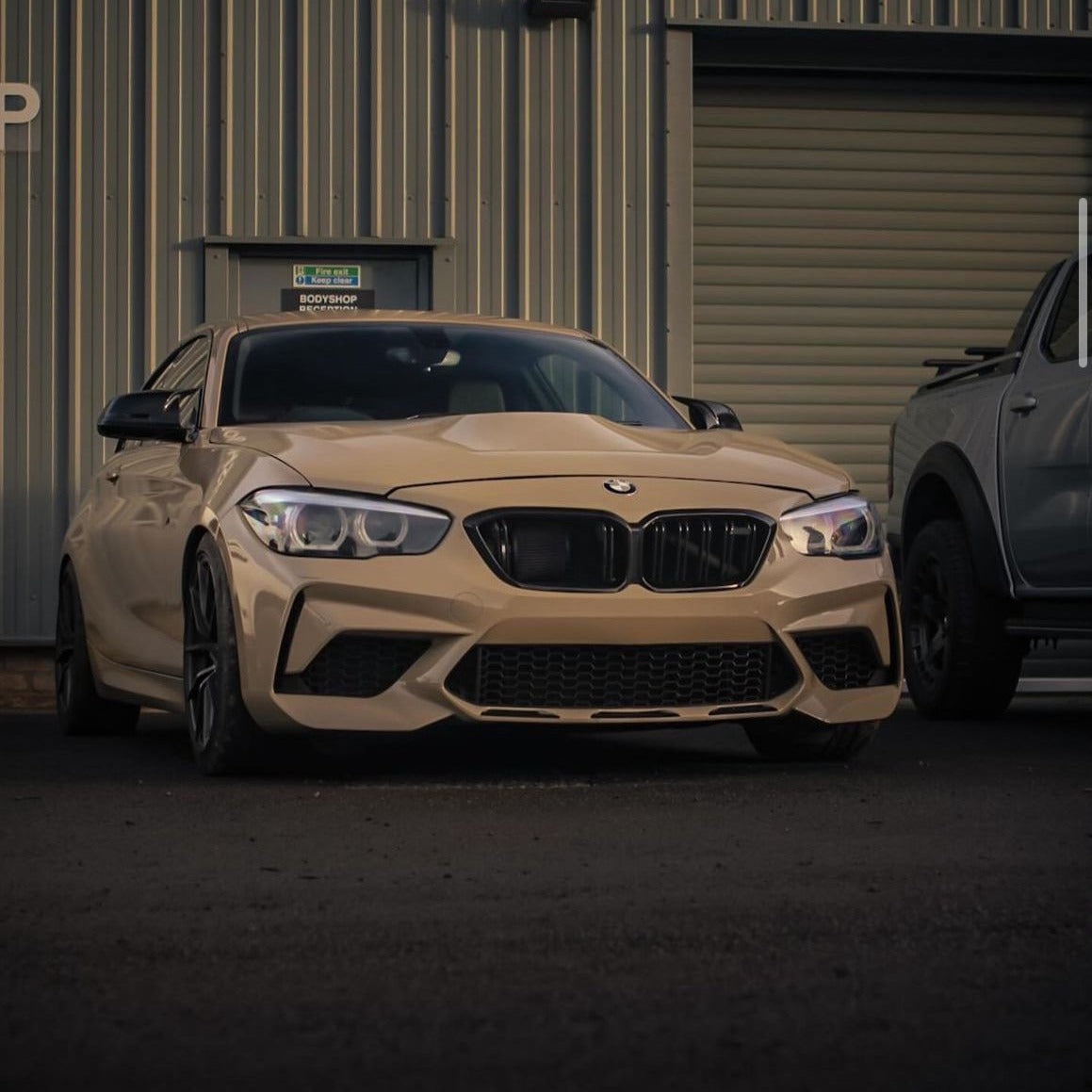 A beige BMW car is parked on asphalt in front of a gray industrial building with a sign reading "Bodyshop Reception" visible above. Another vehicle is partially seen beside it.