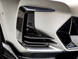 A carbon fiber car bumper features intricate aerodynamic vents and sleek curves, set against a smooth, metallic body, resting on textured asphalt.