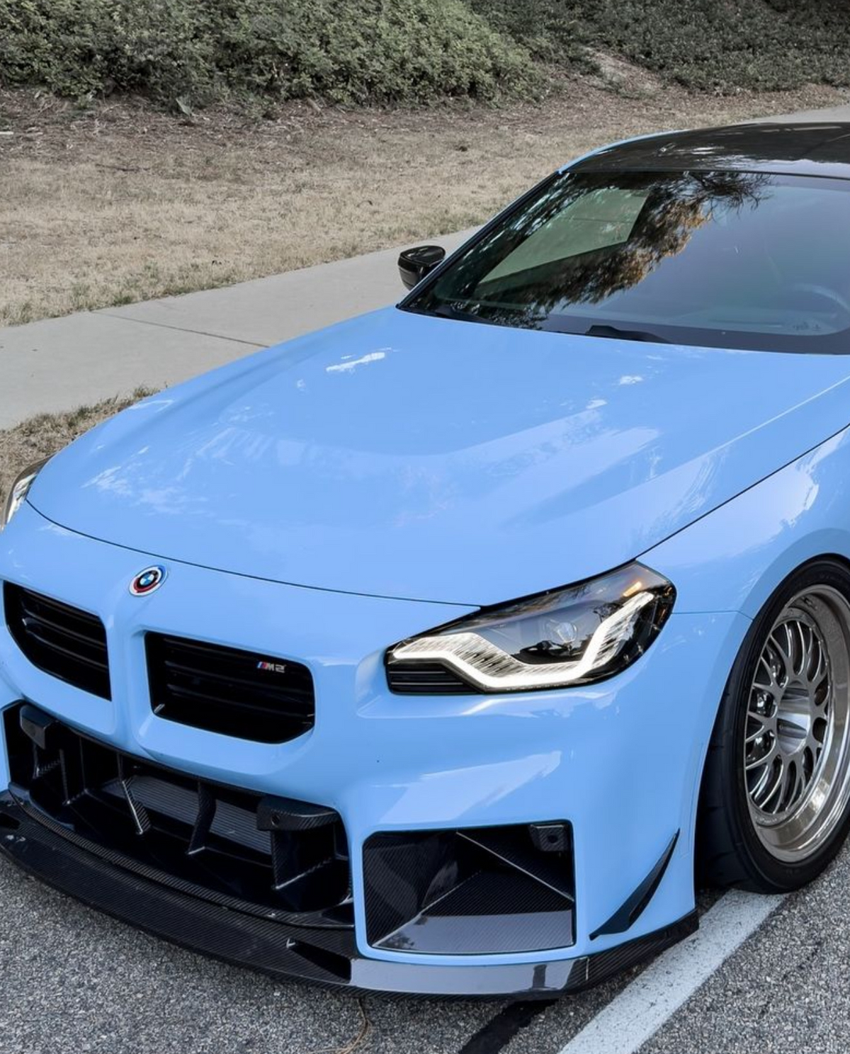 A sleek, blue sports car is parked on a road next to a grassy area, featuring a glossy black roof and distinctive front bumper with an "M2" badge.