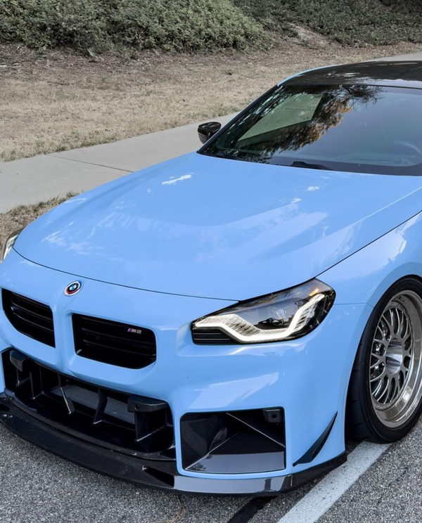 A sleek, blue sports car is parked on a road next to a grassy area, featuring a glossy black roof and distinctive front bumper with an "M2" badge.