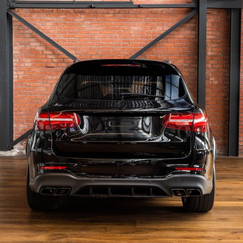 A black SUV is parked indoors on a wooden floor, facing a brick wall with visible metal beams. Other vehicles are partially visible to the sides in the well-lit space.