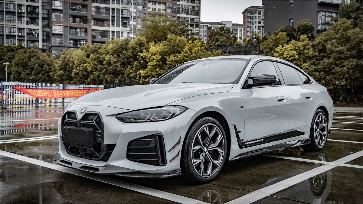 A white sports car is parked on a wet pavement, displaying aerodynamic features. It's surrounded by urban residential buildings and trees. A colorful banner with Chinese characters is in the background.