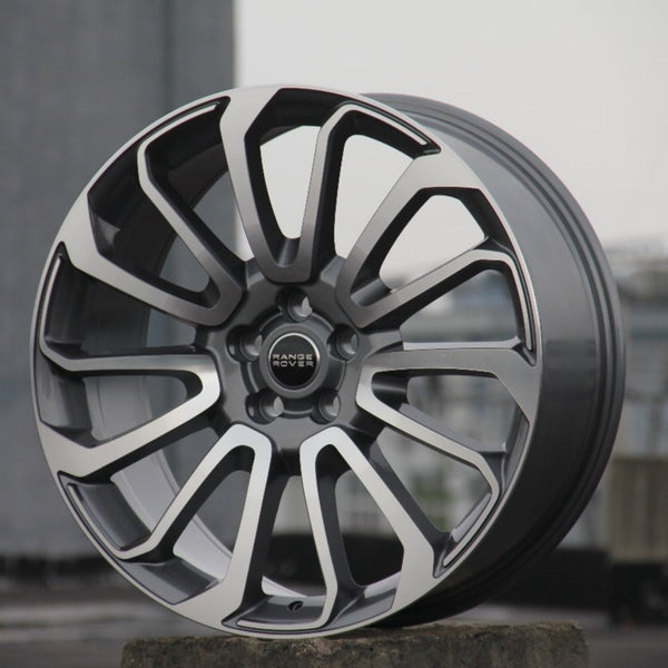 A polished silver and black alloy wheel rests upright on concrete. "Range Rover" is inscribed at its center. The background is blurred, suggesting an urban setting.