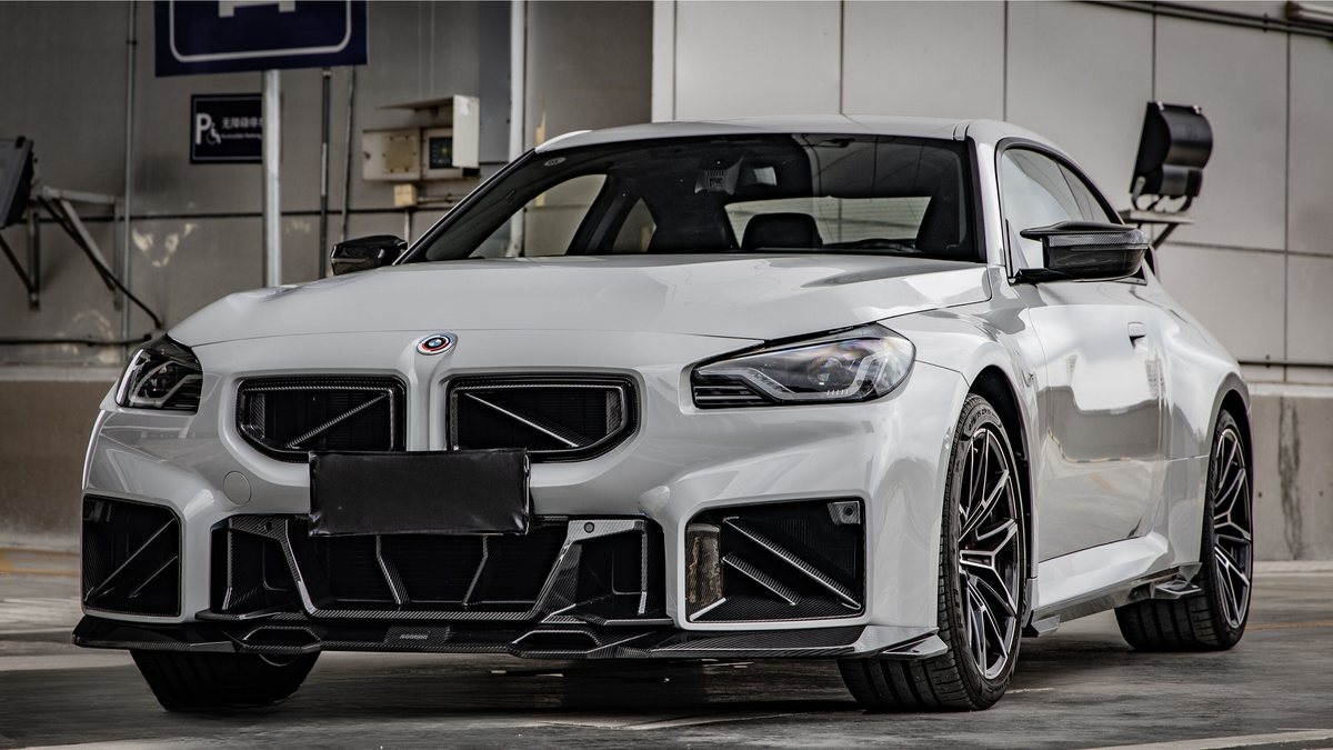 A white sports car is parked in a garage, displaying a sleek body with aggressive front styling. The setting includes a wall-mounted parking sign and exposed industrial elements.