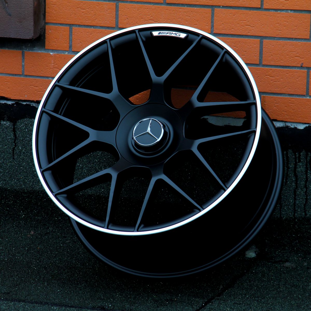 A black alloy wheel with a Mercedes-Benz logo and "AMG" text rests against an orange brick wall on a concrete surface.