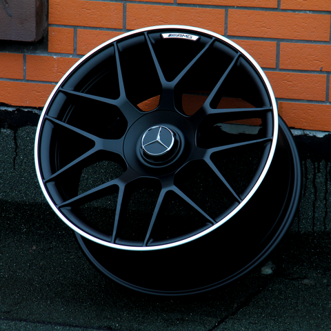 A black alloy wheel with a Mercedes-Benz logo and "AMG" text rests against an orange brick wall on a concrete surface.