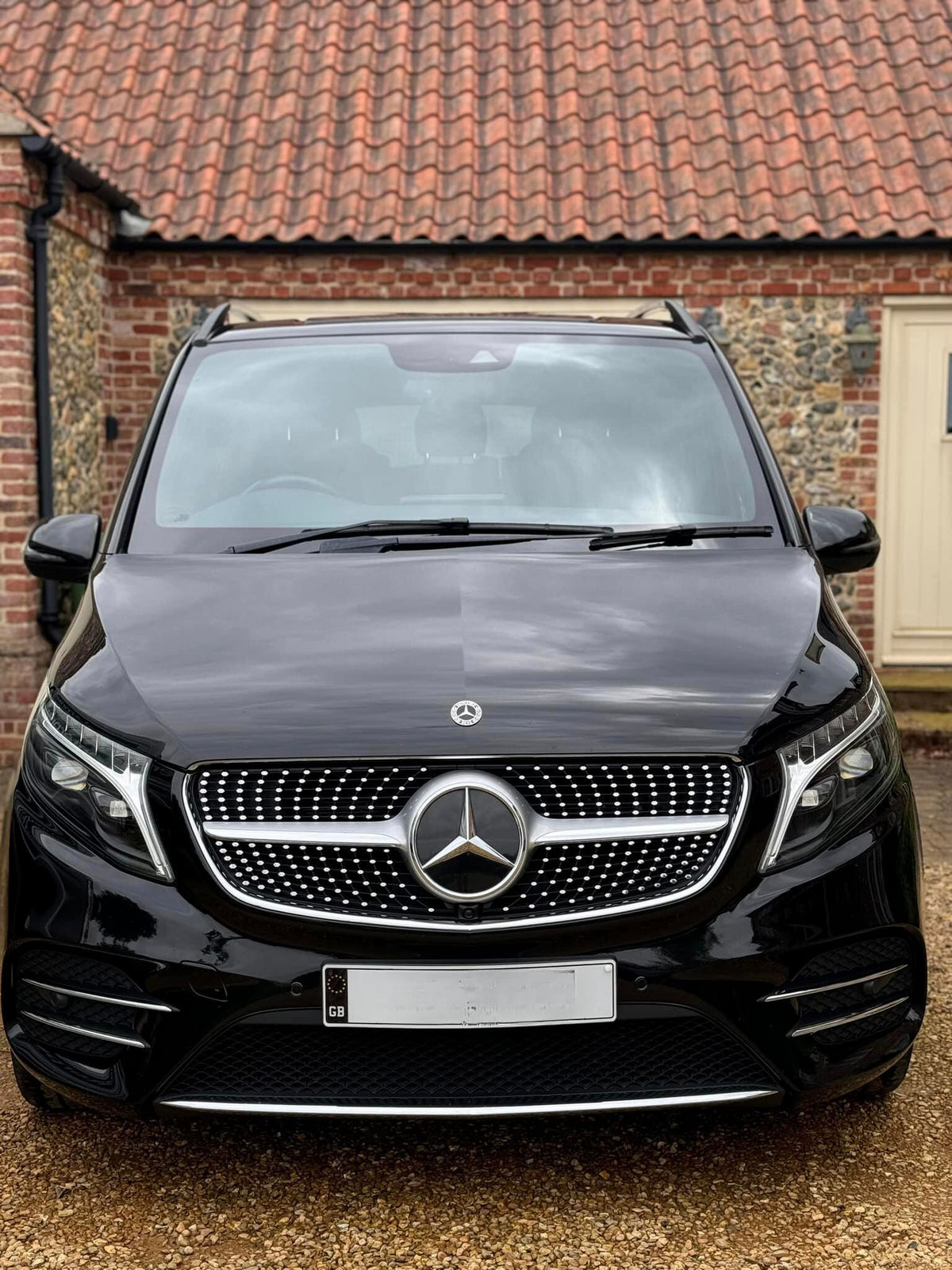 A black Mercedes-Benz van is parked on a gravel driveway in front of a brick building with a tiled roof. The license plate shows a "GB" emblem.