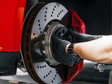 A person's hand, wearing black gloves, installs or inspects a perforated brake disc on a car, with a red caliper, in a garage or workshop setting.