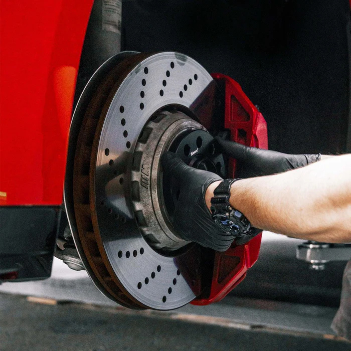 A person's hand, wearing black gloves, installs or inspects a perforated brake disc on a car, with a red caliper, in a garage or workshop setting.