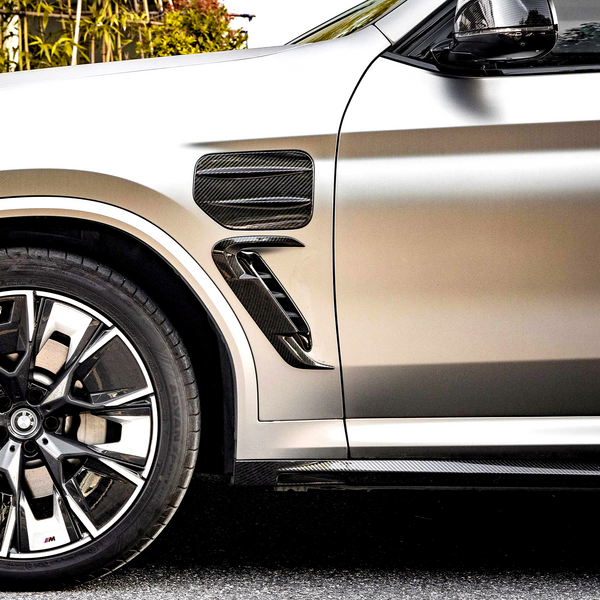 A car’s front side panel displays carbon fiber vents and a shiny metallic finish. The detailed wheel, featuring the BMW logo, occupies the left side. The background has greenery.