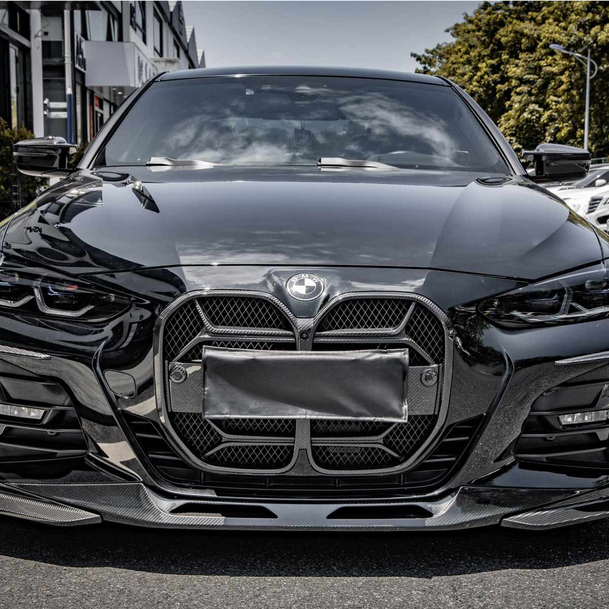 A sleek black car is parked on the side of a street, with modern headlights and a distinctive front grille. Buildings and parked cars line the background.