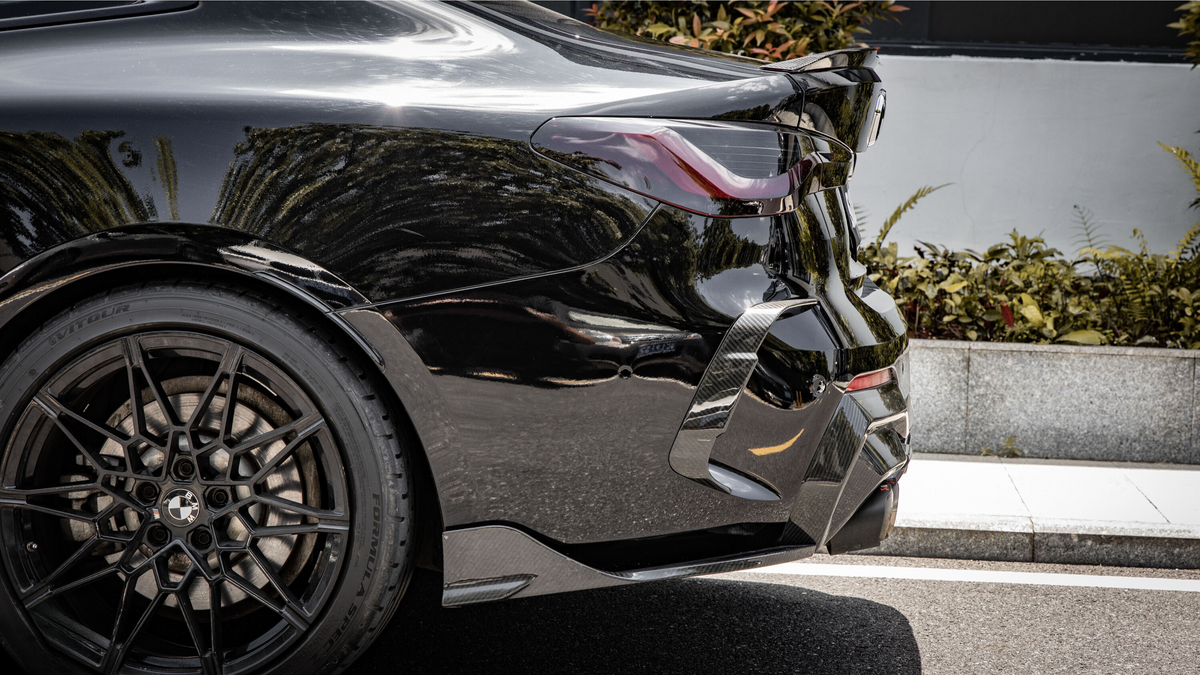 A black car is parked on a street, with sunlight reflecting on its polished surface. It features sleek, curvy design lines and sporty alloy wheels, bordered by greenery and a curb.