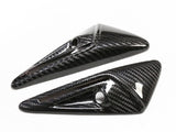 Two glossy, carbon fiber-finished, triangular objects with small circular holes lie angled on a white surface.