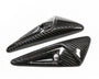 Two glossy, carbon fiber-finished, triangular objects with small circular holes lie angled on a white surface.