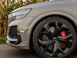 A sleek car front with a black alloy wheel and red brake caliper rests on a paved road, surrounded by tall corn plants. The tire reads 