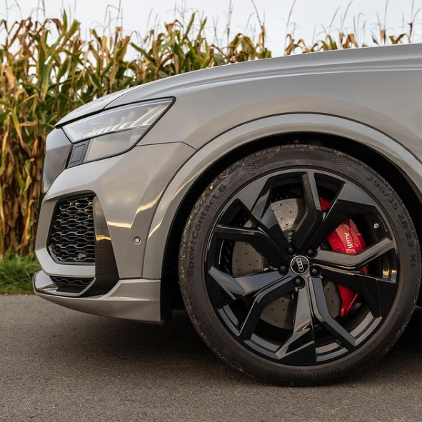 A sleek car front with a black alloy wheel and red brake caliper rests on a paved road, surrounded by tall corn plants. The tire reads "SportContact 6 285/35 ZR 22 XL."