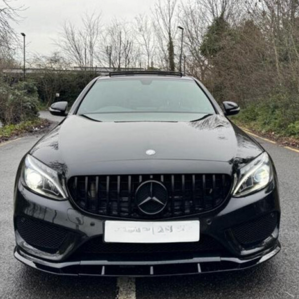 A black Mercedes car is parked on a road lined with leafless trees and bushes. It has illuminated headlights and a visible Mercedes emblem at the front center.