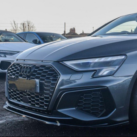 A grey Audi car is parked on a gravel lot. Its front grille is prominent, adjacent to another white car. A fence and trees are visible in the background.