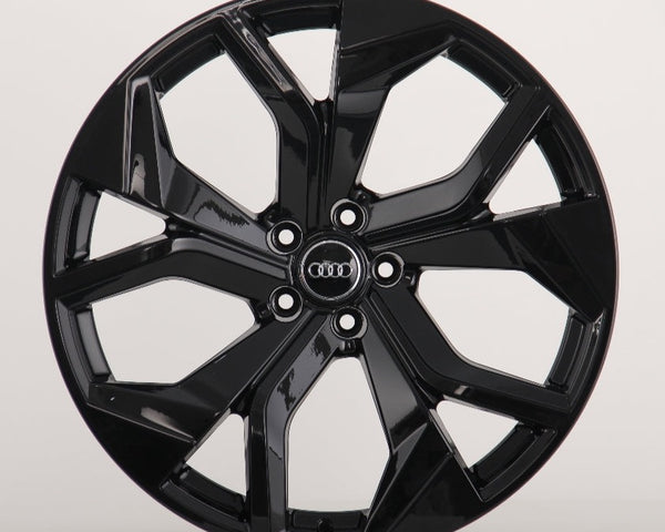 A glossy black car wheel features a five-spoke design with intersecting shapes, prominently displaying the Audi logo at the center on a plain, light background.