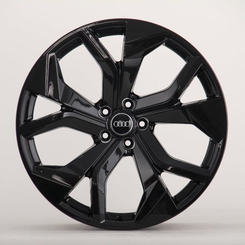 A glossy black car wheel features a five-spoke design with intersecting shapes, prominently displaying the Audi logo at the center on a plain, light background.