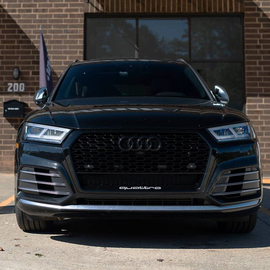 A black Audi SUV, featuring the logo and "quattro," is parked in front of a brick building. The number "200" is visible on the wall.