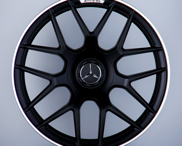 A black Mercedes-AMG wheel with a multi-spoke design stands upright against a gray background. The Mercedes logo is centered, with "AMG" text at the top of the wheel.