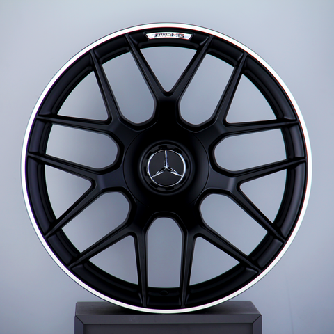 A black Mercedes-AMG wheel with a multi-spoke design stands upright against a gray background. The Mercedes logo is centered, with "AMG" text at the top of the wheel.