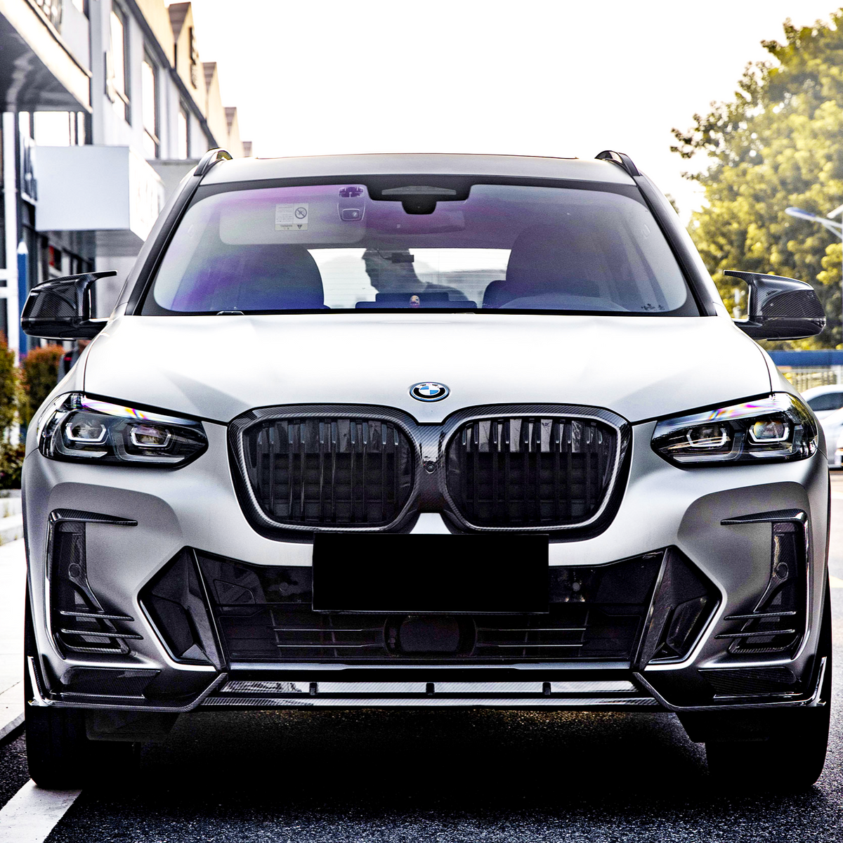 A sleek, white SUV with a prominent grille is parked on a street. It has tinted windows and modern LED headlights, with buildings and greenery in the background.