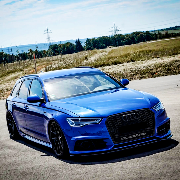 A blue Audi station wagon, parked on a road, features black wheels and a "quattro" badge on the grille. It's surrounded by fields and distant trees under a clear sky.
