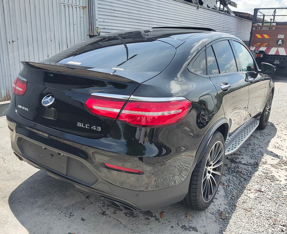 A black car labeled "GLC 43" and "AMG" is parked on gravel beside a corrugated metal wall and a truck. Bright sunlight casts shadows on the ground.