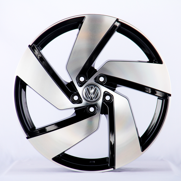 A sleek, silver and black alloy wheel with an intricate design sits upright in a well-lit studio. The central emblem features the Volkswagen logo against a plain white background.