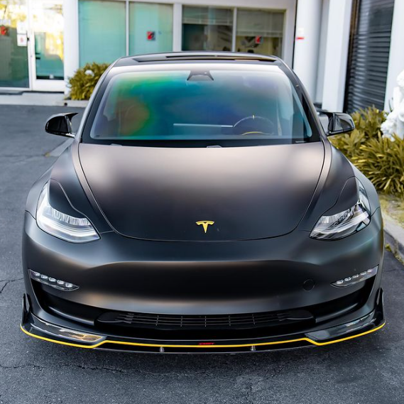 Car facing forward, black matte finish, with yellow accents and Tesla logo visible. Parked on an asphalt surface, flanked by green plants and modern white building in the background.