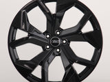 A glossy black car wheel rim with five split spokes, featuring the Audi logo in the center, is displayed against a plain white background.