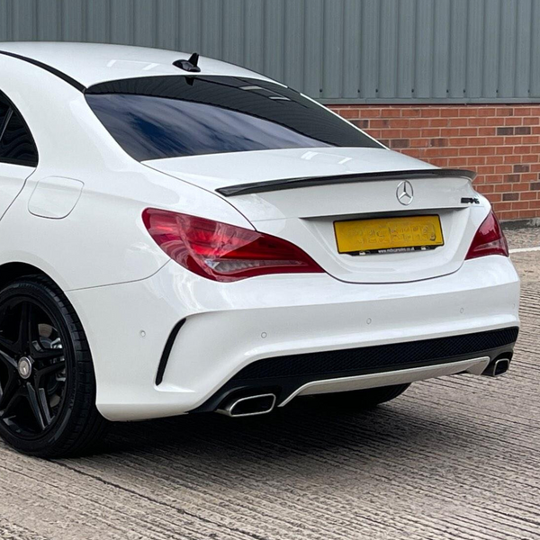White car parked on a concrete surface, featuring tinted windows, a rear spoiler, and dual exhausts. The background shows a gray corrugated metal wall with a lower brick section.