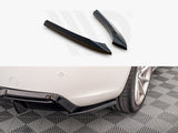 Two black car diffuser fins rest on a white surface above a close-up of a car's rear bumper with a diffuser installed, parked on a patterned grey floor.
